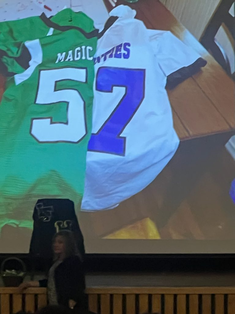 Dorothy Connor speaking at assembly with image in background of overlapping Herkimer Magicians and Little Falls Mounties football jerseys.