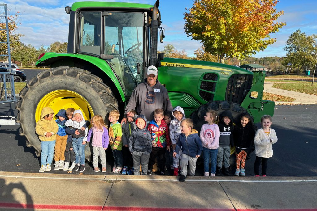 Preschool students gather with an adult in front of a giant tractor during a fall outing.