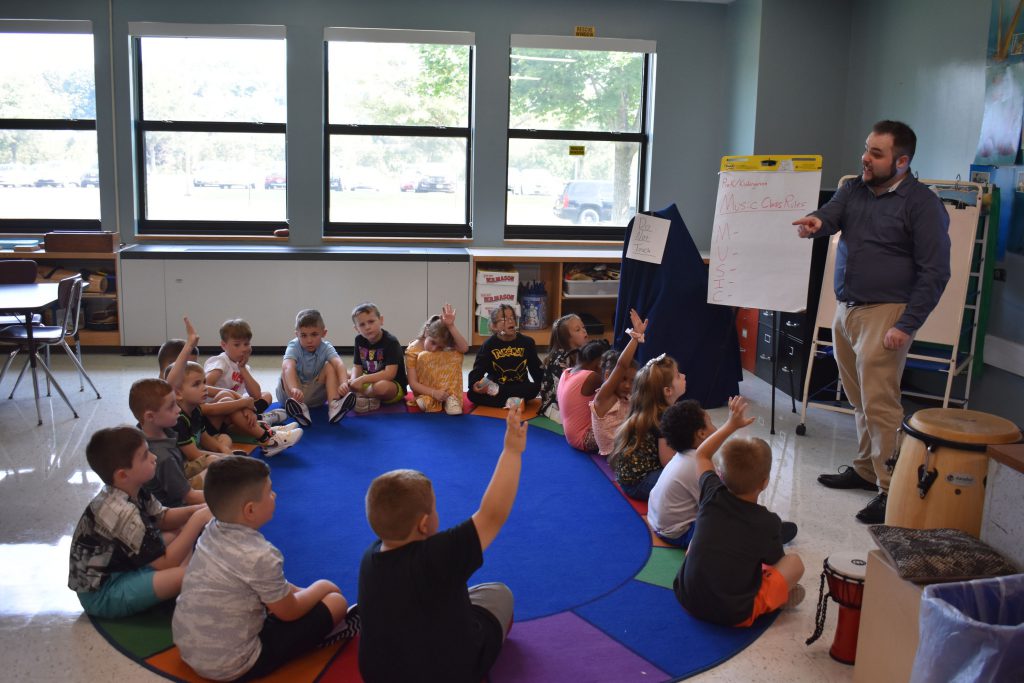 Elementary students sit on the floor and raise hands while the teacher presents a question.