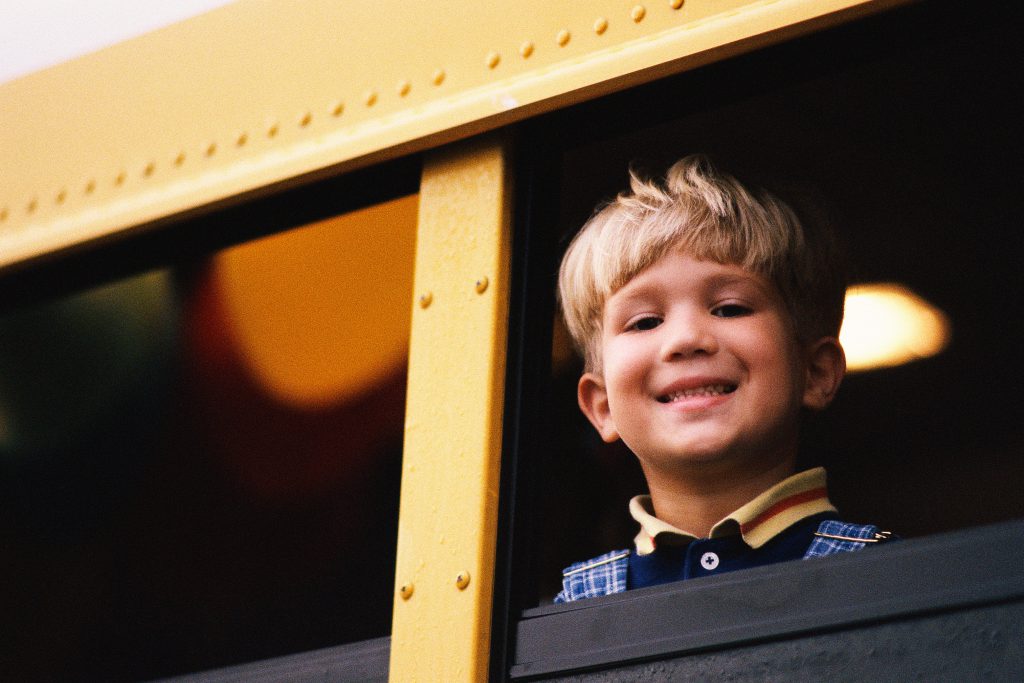 A elementary aged child smiles from a school bus window.