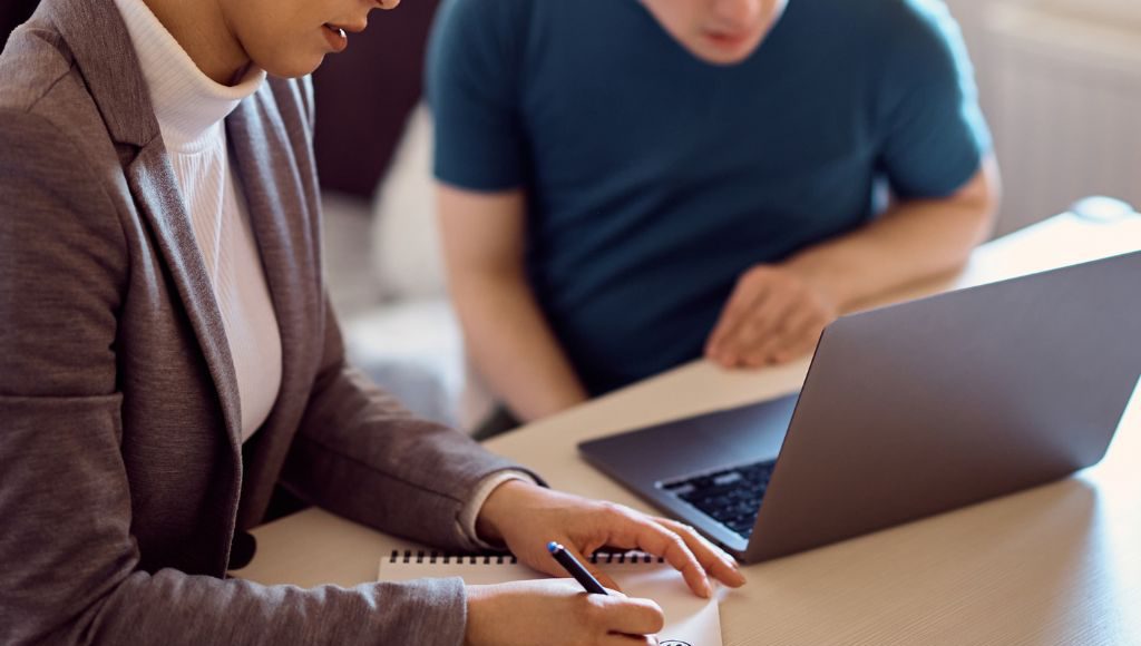 An adult writes notes while a high schooler peers onto a laptop screen.