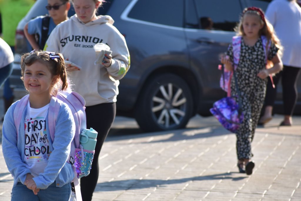 An elementary student arrives to school wearing a t-shirt that says "First Day of School"