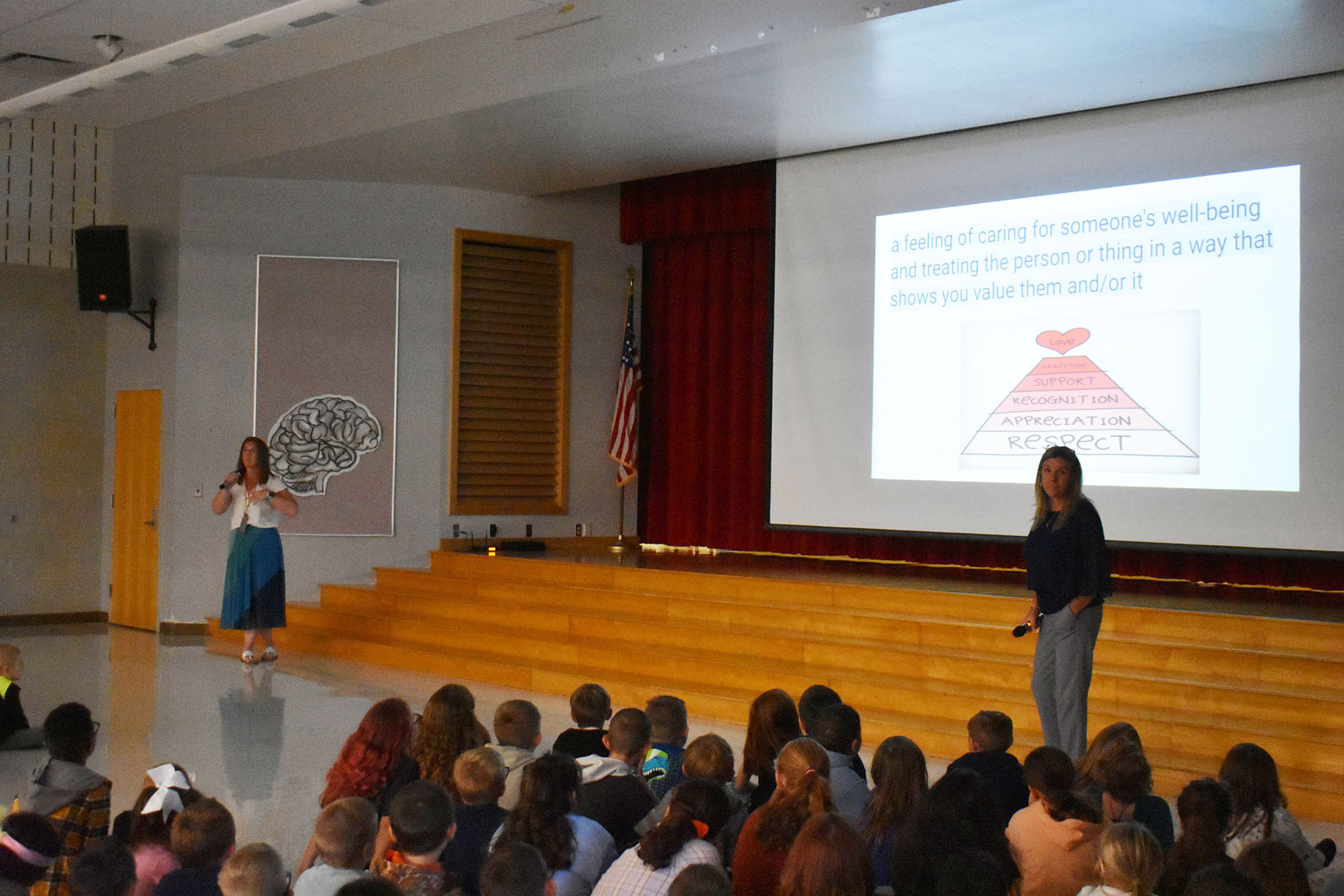 Principal and teacher speaking in front of students in cafeteria auditorium