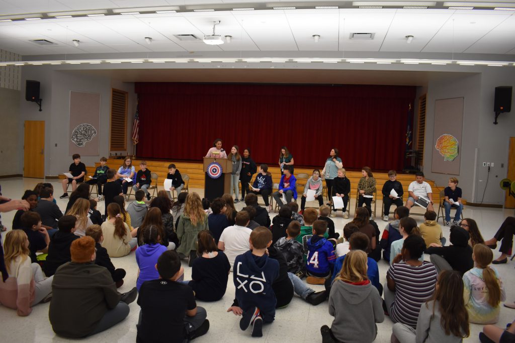 Fifth graders taking part in an election event in cafeteria. Candidates and hosts up front with other students looking on.