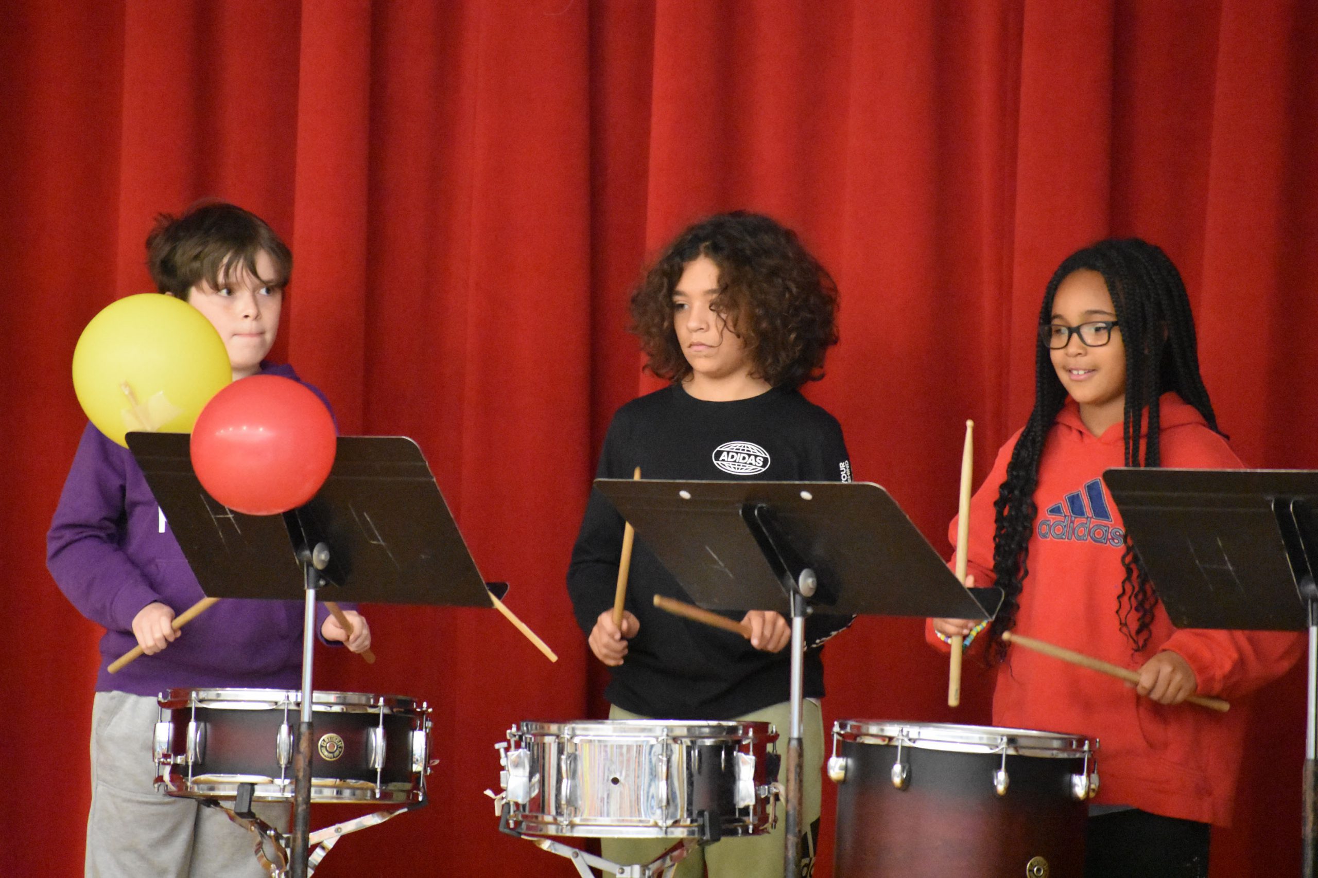 Three students playing drums on stage in front of red curtain